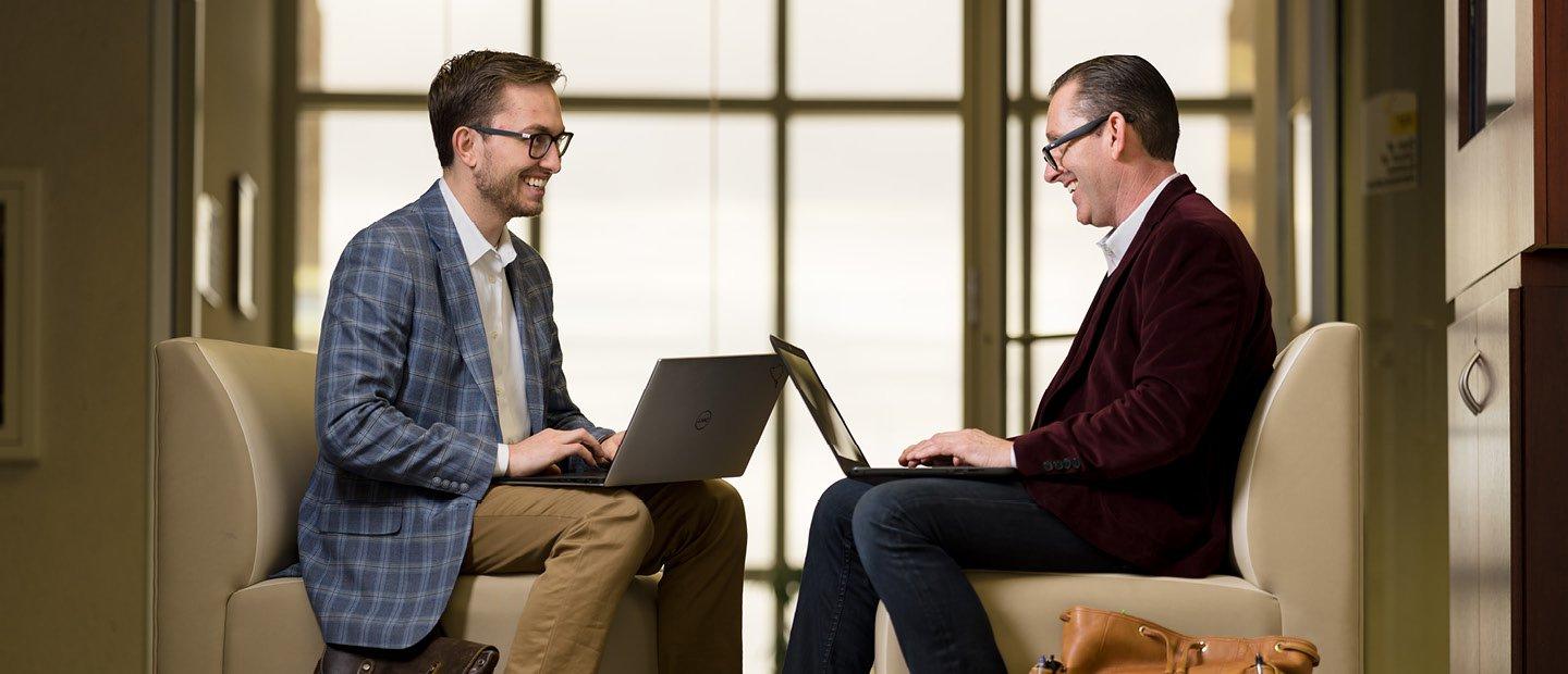 Two men seated in leather chairs with laptops, facing each other.